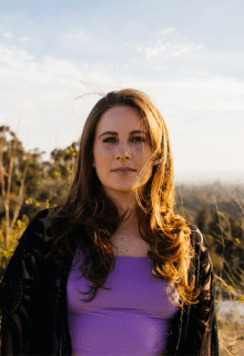 A woman with long brown hair and a purple top stands outdoors with trees and sky in the background.