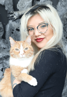 A woman with blonde hair and glasses holds an orange and white cat, both facing the camera, with a gray textured background behind them.