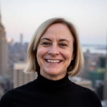 A smiling woman with blonde hair wearing a black turtleneck in front of a city skyline.